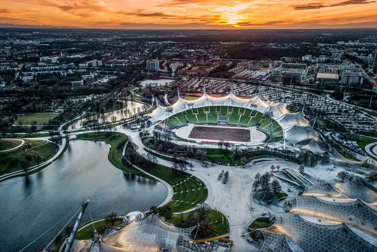 A European city seen from the air at sunset, a lake and parkland in the foreground beneath the sweeping tented roofs of a stadium complex, with the built-up skyline running to the horizon: the destination side of the move, and what the cessation date and the transfer allowances have to be sequenced around