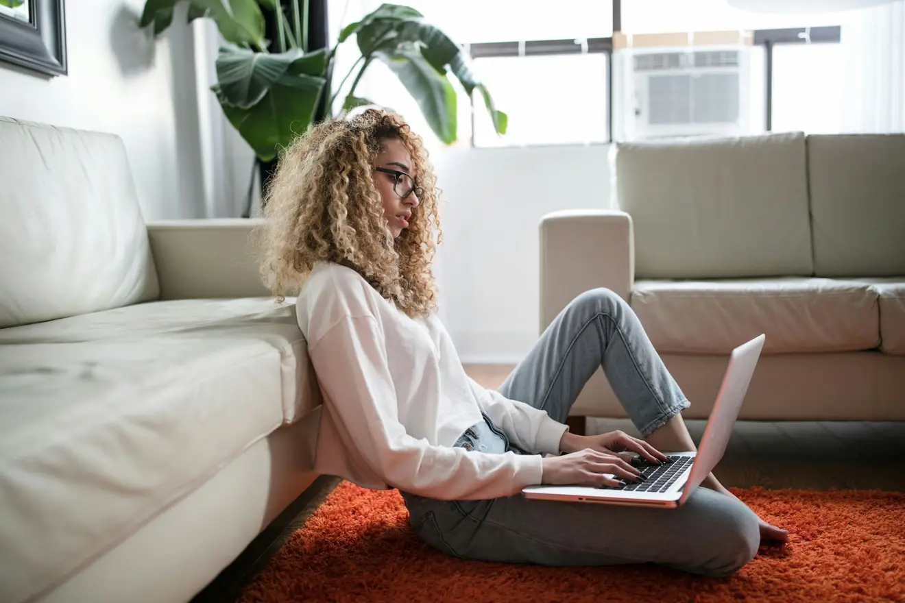 A woman sitting on a living room floor with her back against a sofa, typing on a laptop balanced on her knees with a large plant and a bright window behind her, the everyday setup the expanded OECD Commentary now asks whether to treat as a fixed place of business