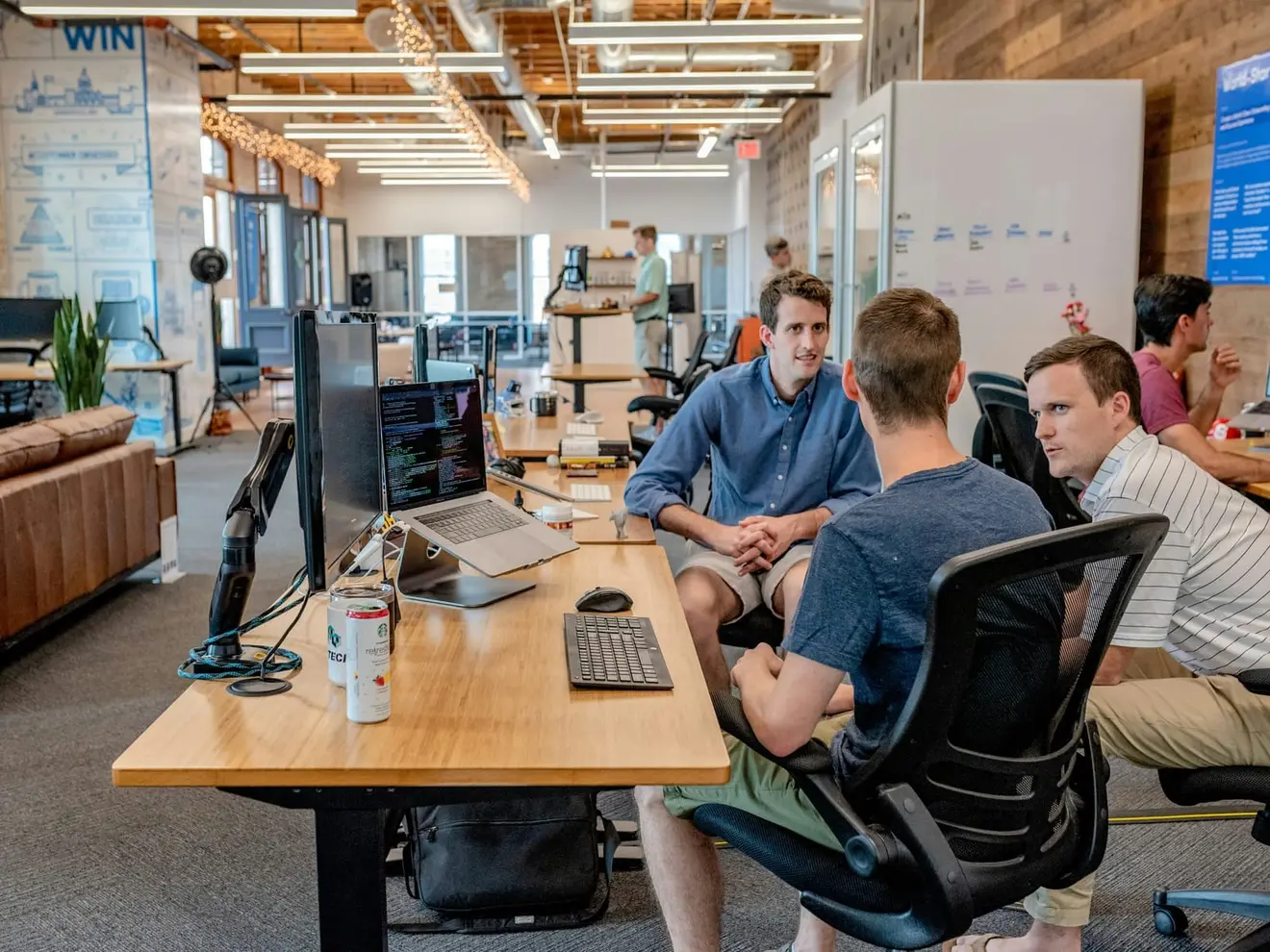 Four people talking around a long wooden desk in an open-plan office, a laptop and external monitors in the foreground and whiteboards on the wall behind.