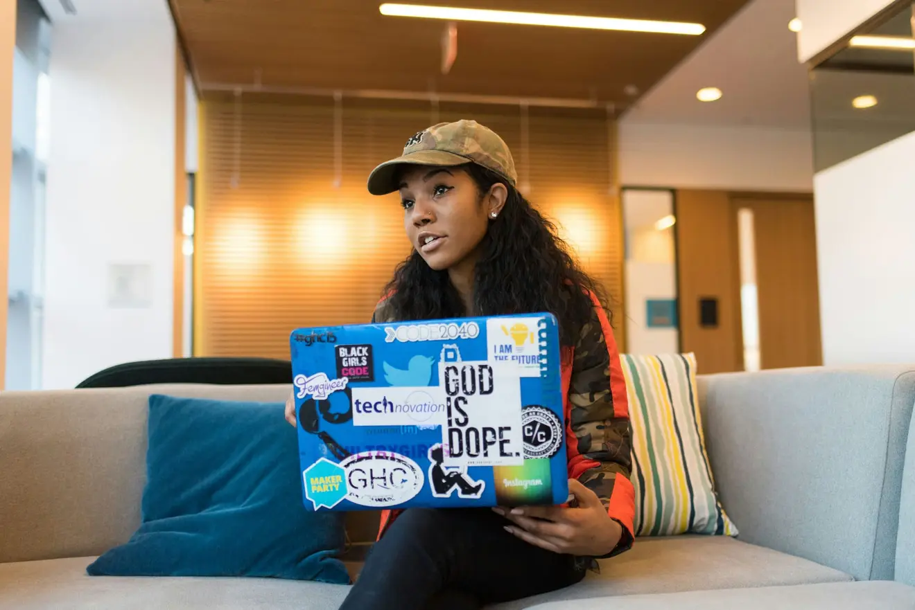 A young founder in a camouflage cap sitting on a sofa in an office lounge, holding a laptop covered in tech and community stickers while talking to someone off camera