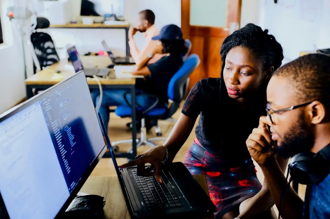 A woman points at a laptop keyboard while a colleague in glasses with headphones round his neck watches the screen, two more people working at a desk behind them.