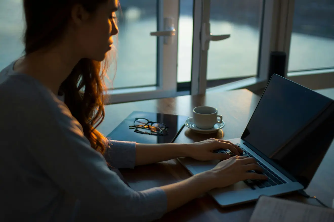 A woman working alone at a laptop by a large window at dusk, with reading glasses and a cup of coffee on the desk beside her