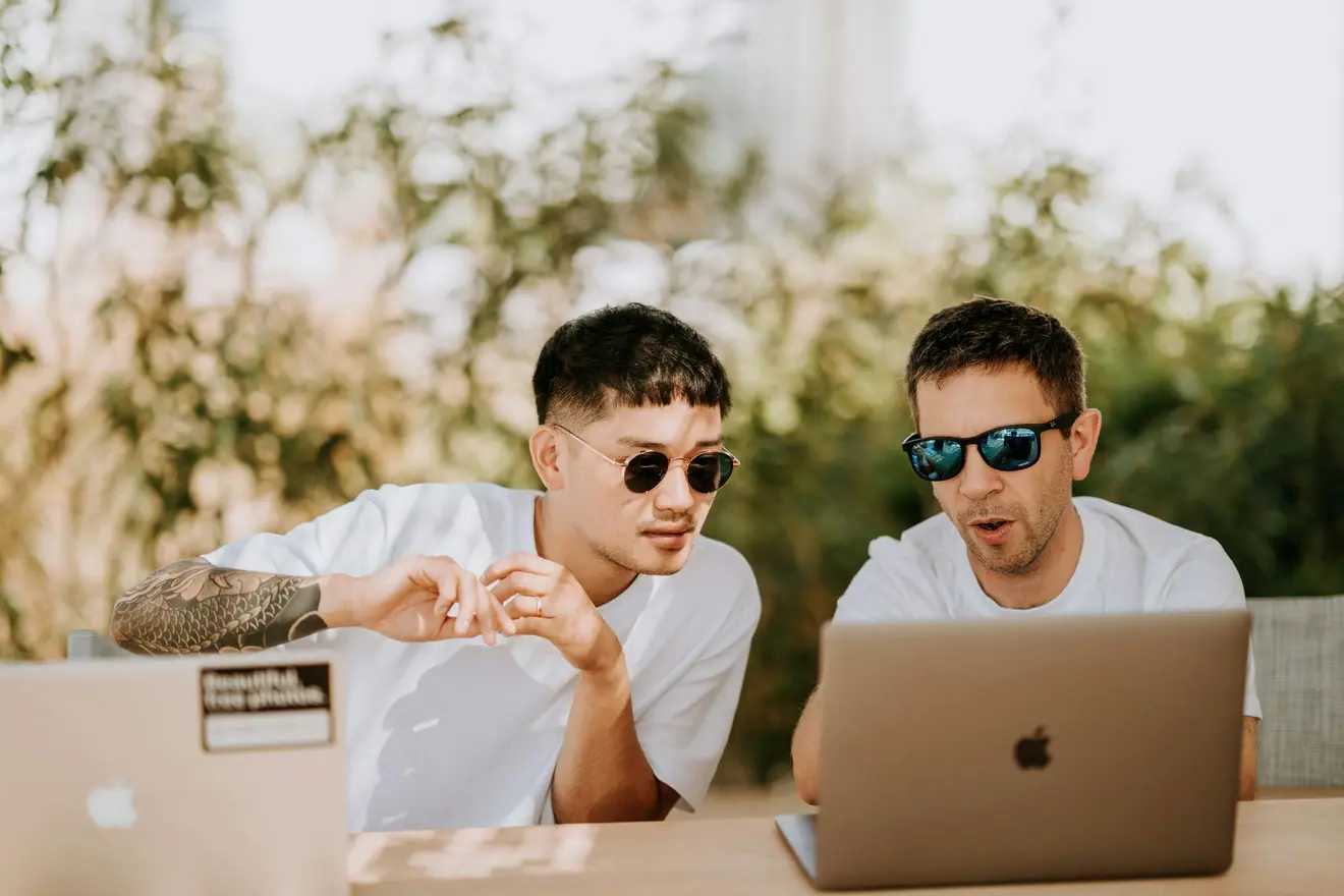 Two men in white t-shirts and sunglasses sitting outdoors at a wooden table with two open laptops, one of them reacting to something on the screen with his mouth open. Founders pricing an exit or a few dividend-heavy years abroad tend to have that reaction to the temporary-non-residence arithmetic
