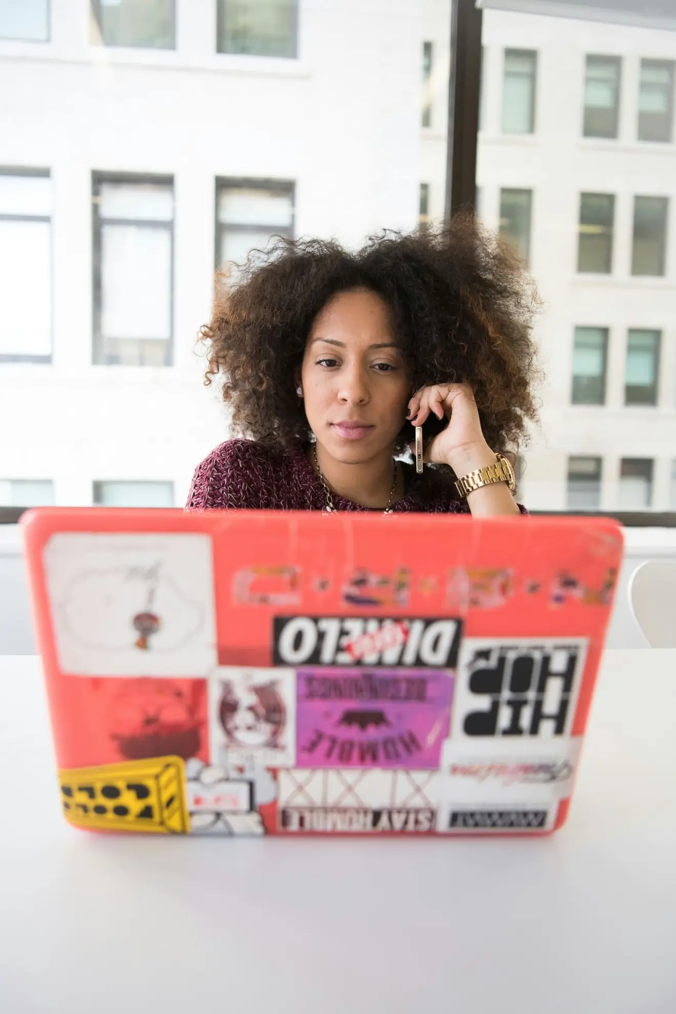 A founder taking a call at a laptop covered in stickers, in an office with city buildings framed in the window behind her