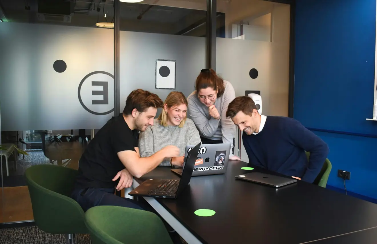 Four colleagues gathered around a table in a glass-walled office, leaning in and laughing at an open laptop between them.