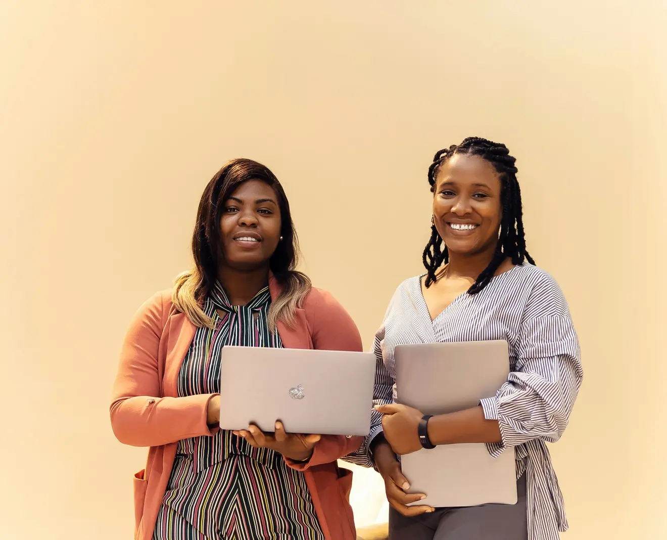 Two women in office clothing standing side by side against a plain warm background, each holding a closed laptop, the kind of local team an employer of record puts a relocating founder's payroll through